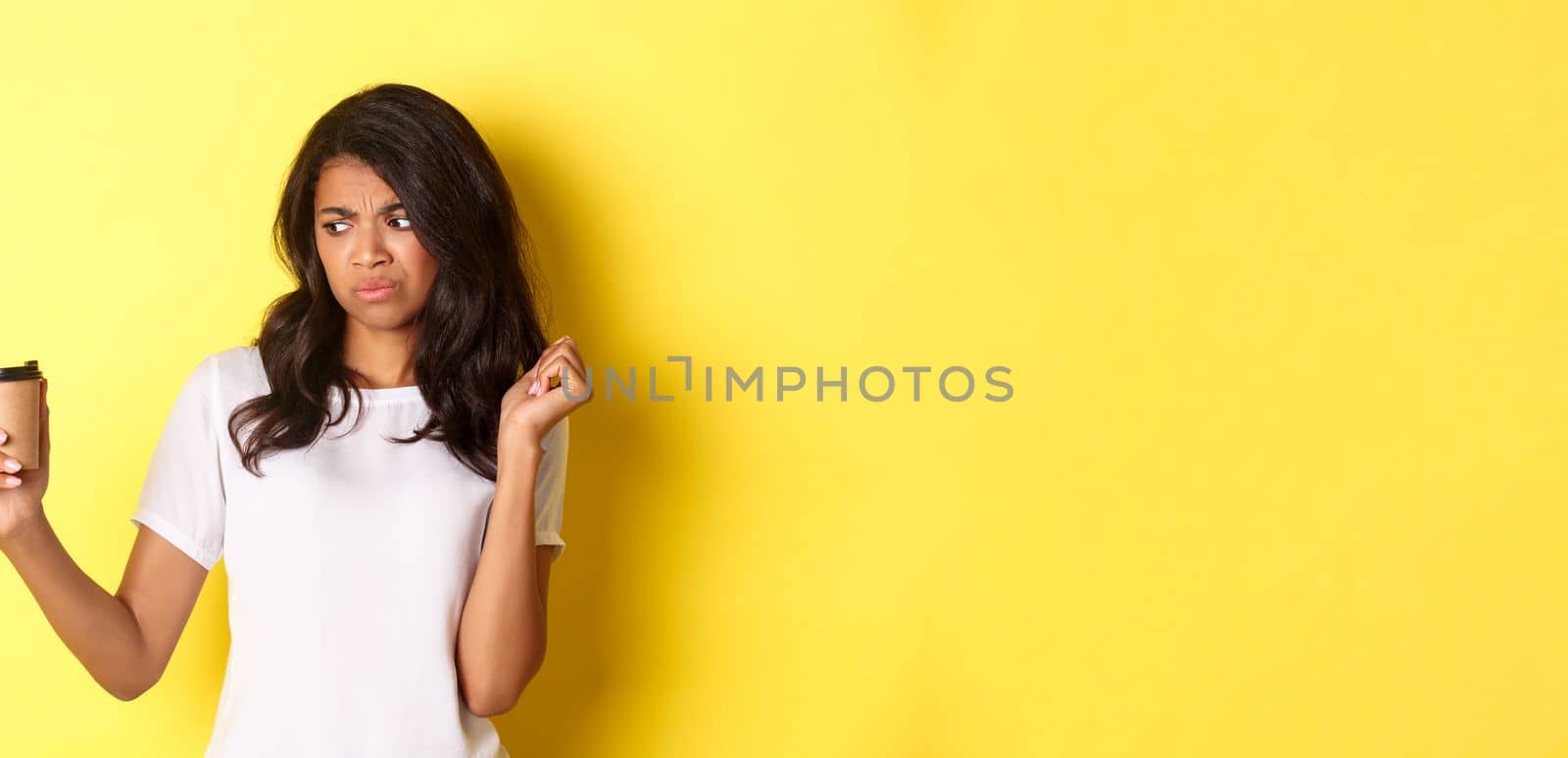 Portrait of african-american girl looking disappointed at cup, dont like bad coffee, standing over yellow background by Benzoix