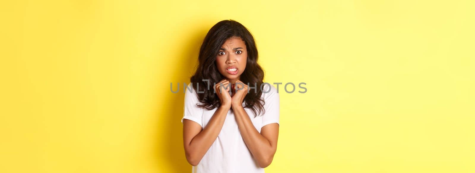 Portrait of cute african-american girl looking scared and reluctant, grimacing displeased, standing over yellow background.