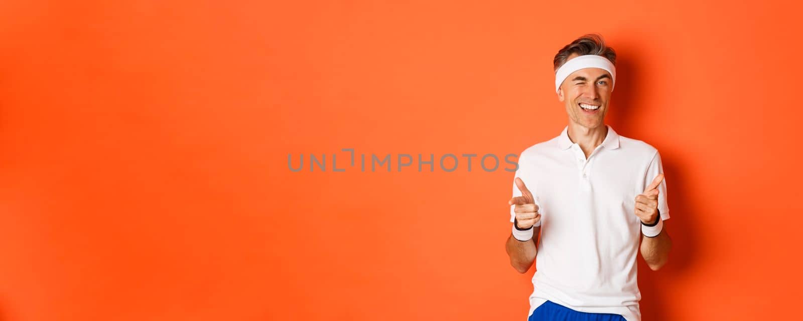 Concept of sport, fitness and lifestyle. Portrait of confident, handsome middle-aged man in workout uniform, pointing fingers at camera and smiling, inviting to gym, standing over orange background.