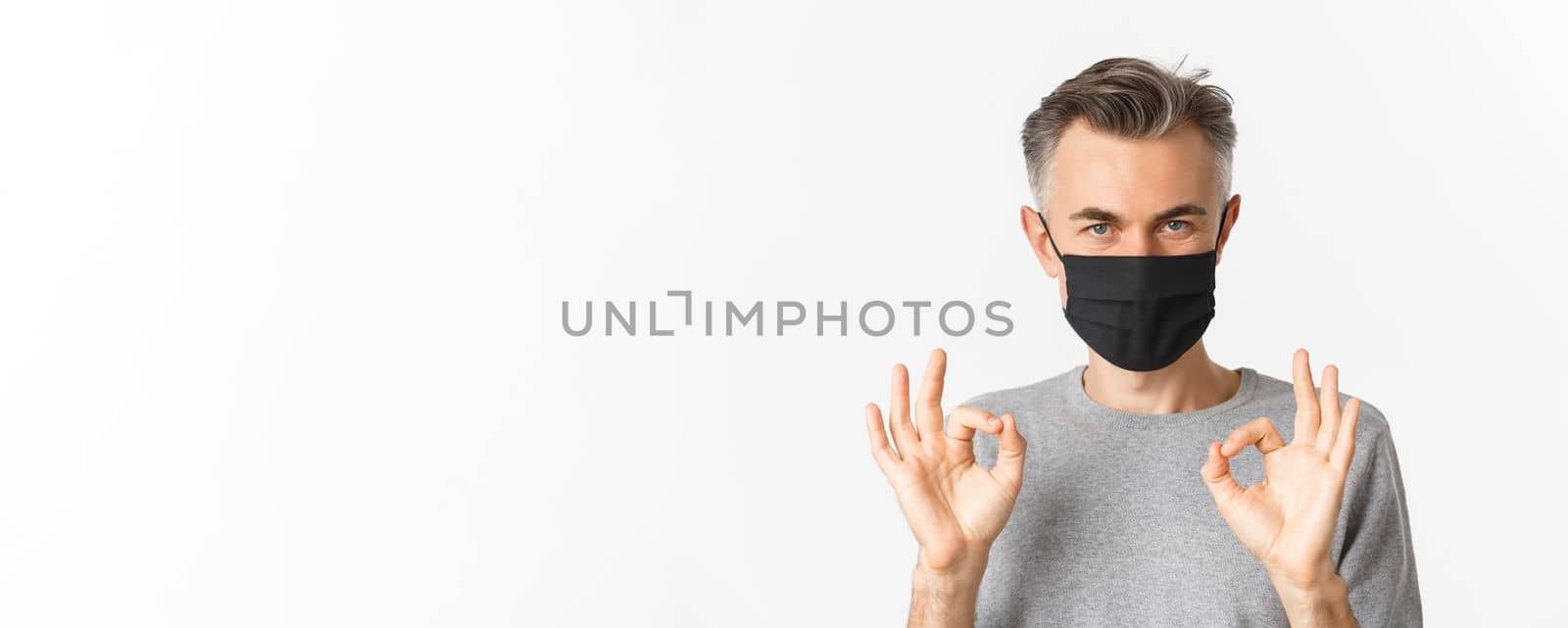 Concept of covid-19, social distancing and quarantine. Close-up of confident middle-aged man, wearing black medical mask, assure everything good, showing okay signs, white background by Benzoix