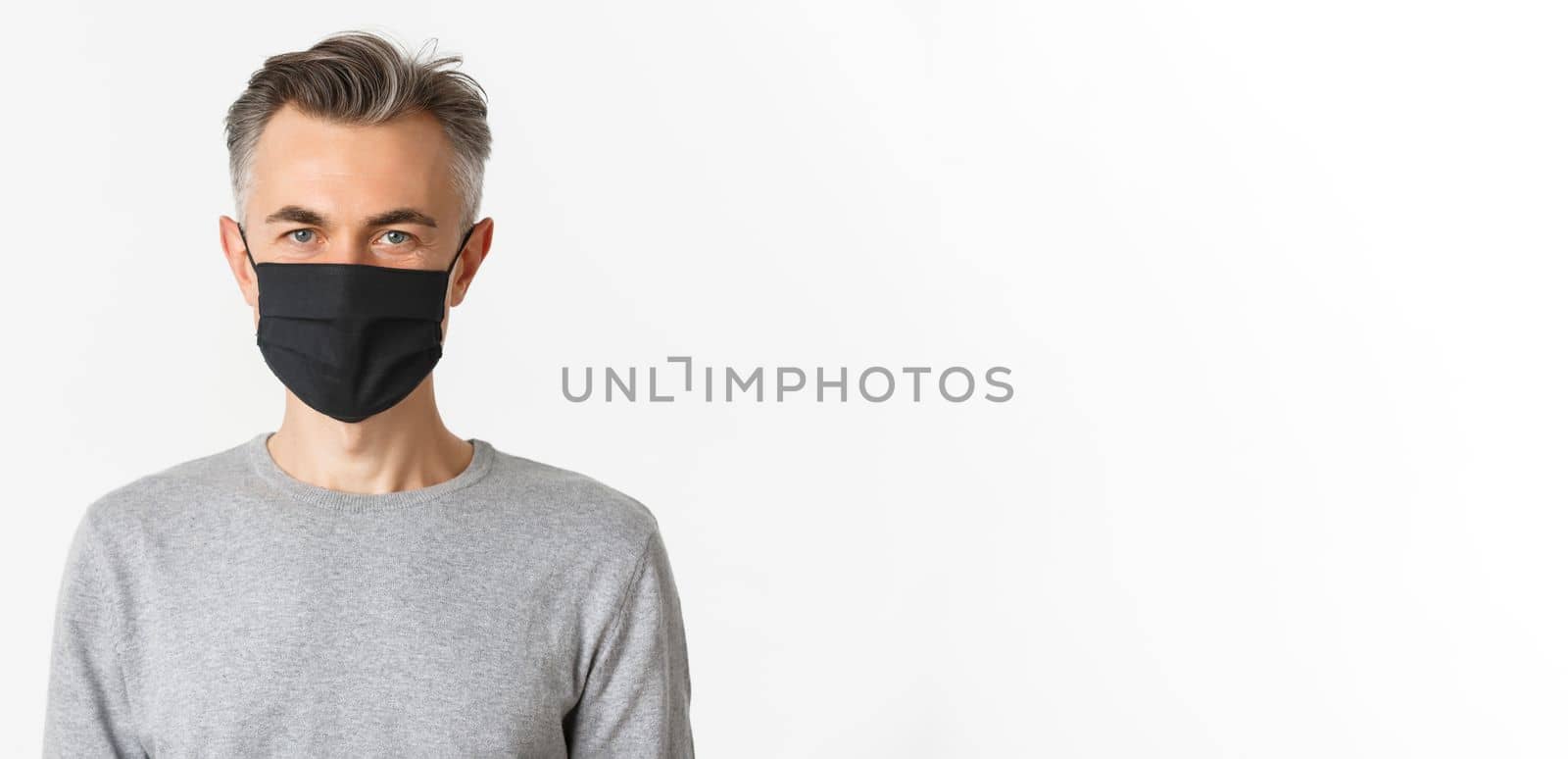 Concept of covid-19, social distancing and lifestyle. Close-up of handsome middle-aged man in black medical mask, looking confident at camera, standing over white background.