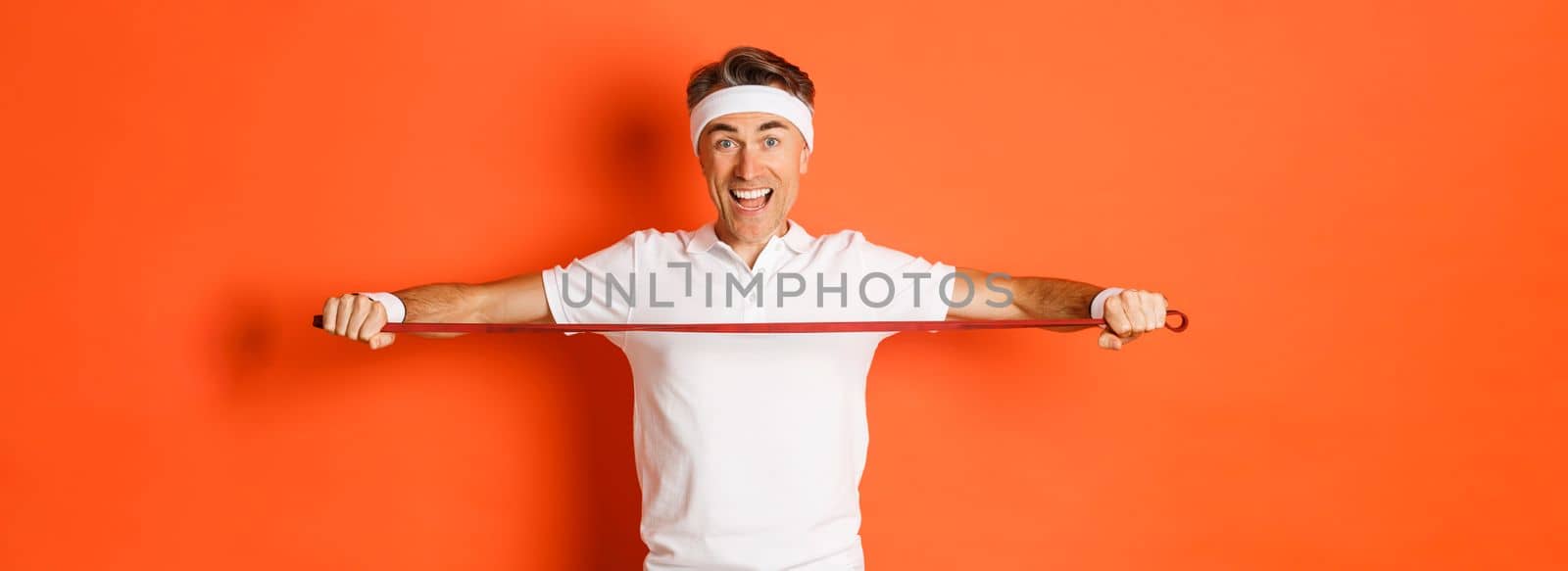 Concept of workout, gym and lifestyle. Active and healthy middle-aged fitness guy, holding stretching rope for exercises and smiling, standing over orange background by Benzoix