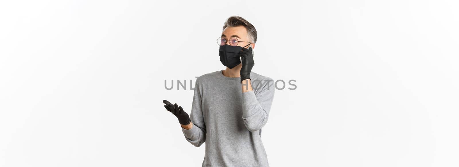 Concept of covid-19, social distancing and lifestyle. Image of handsome middle-aged man in medical mask and gloves, talking on mobile phone, standing over white background.