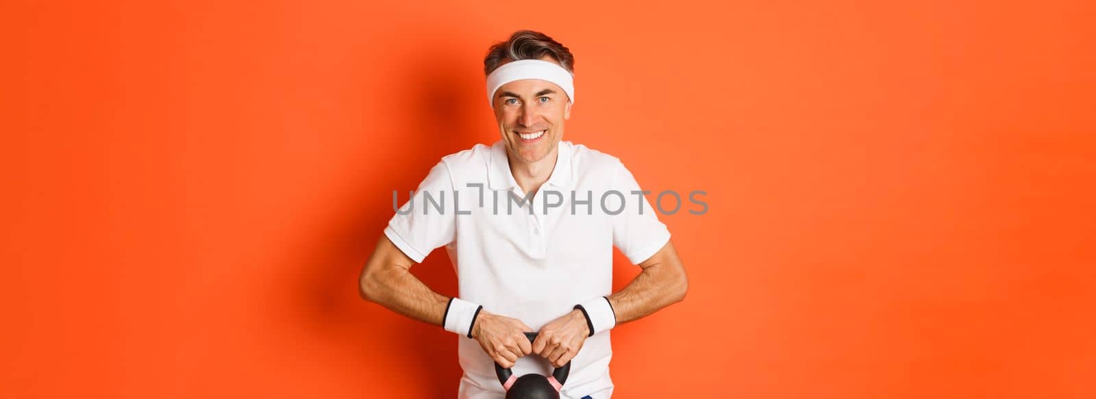 Concept of workout, gym and lifestyle. Image of handsome middle-aged guy doing sport exercises, lifting kettlebell and smiling, standing over orange background.