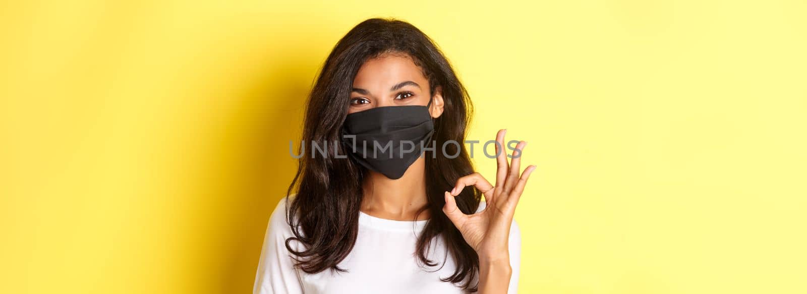 Concept of coronavirus, pandemic and lifestyle. Close-up of pretty african-american woman in black face mask, showing okay sign in approval, praise good work, yellow background by Benzoix