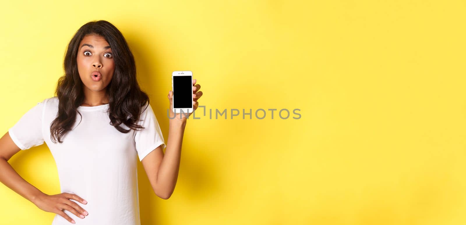 Image of amazed african-american girl, looking fascinated and showing smartphone screen, standing over yellow background.