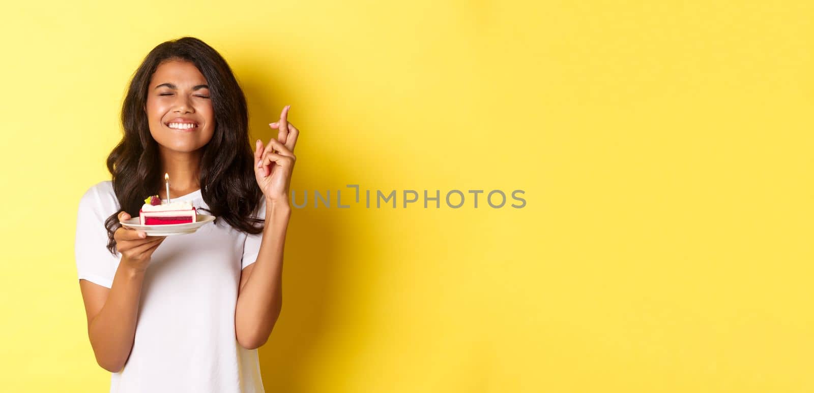 Portrait of cute african-american woman, close eyes and smiling, crossing fingers to make wish on birthday cake, celebrating b-day, standing over yellow background.
