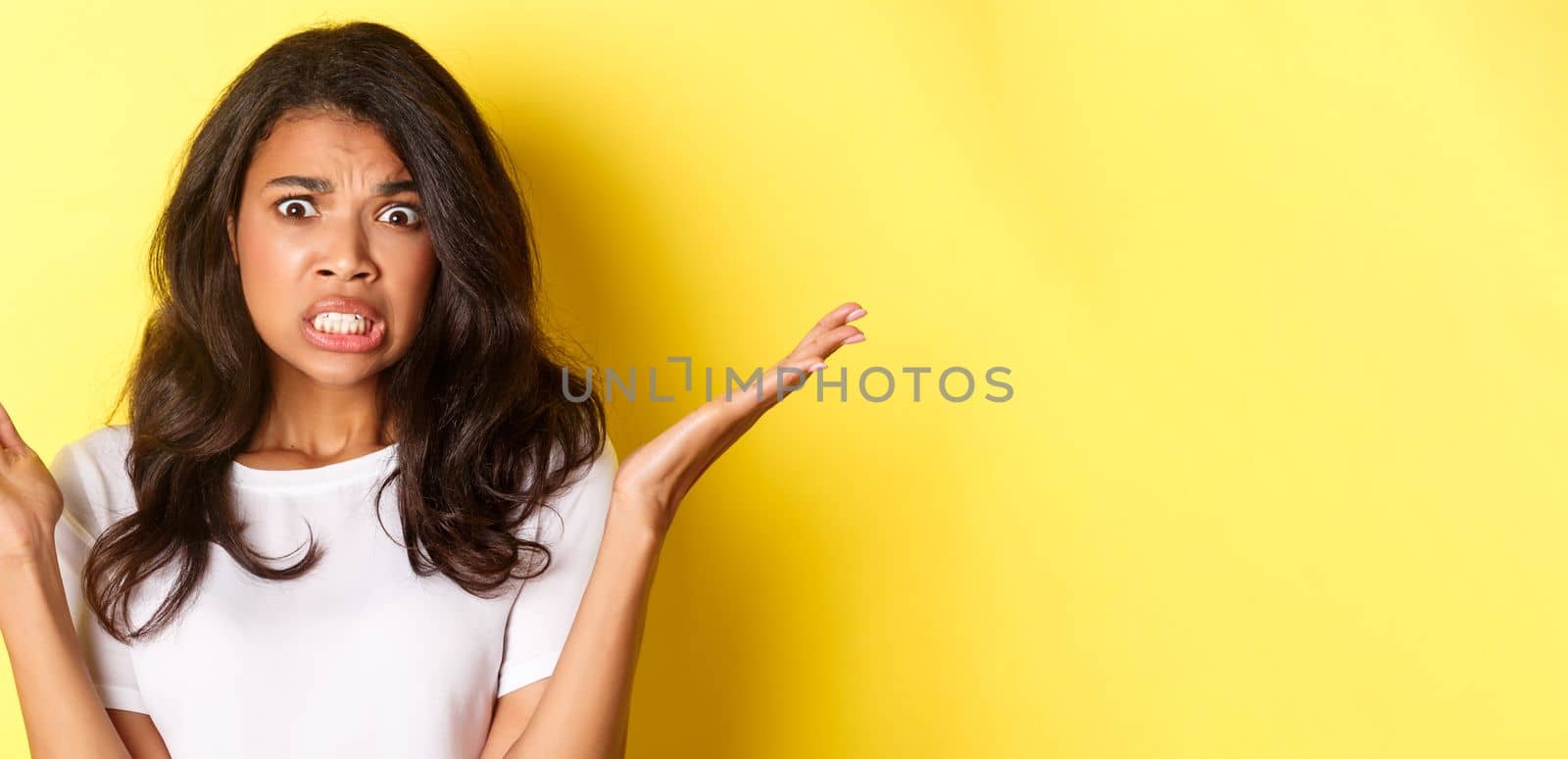 Close-up of frustrated and shocked african-american girl, raising hands up and looking at disaster, standing over yellow background.