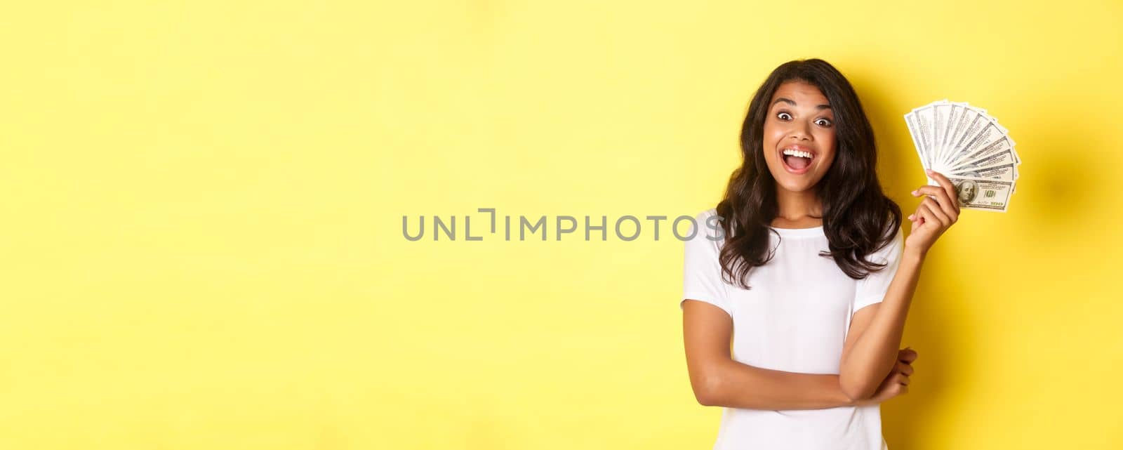 Portrait of beautiful african-american girl, smiling happy and showing money, going shopping, standing over yellow background.