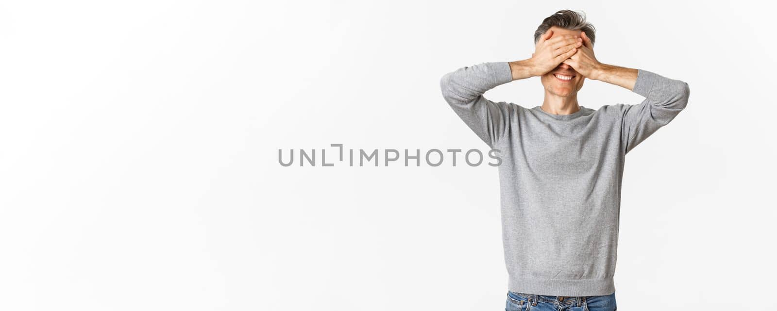 Portrait of handsome happy middle-aged man waiting for surprise, shut eyes and smiling, playing hide n seek, standing over white background.