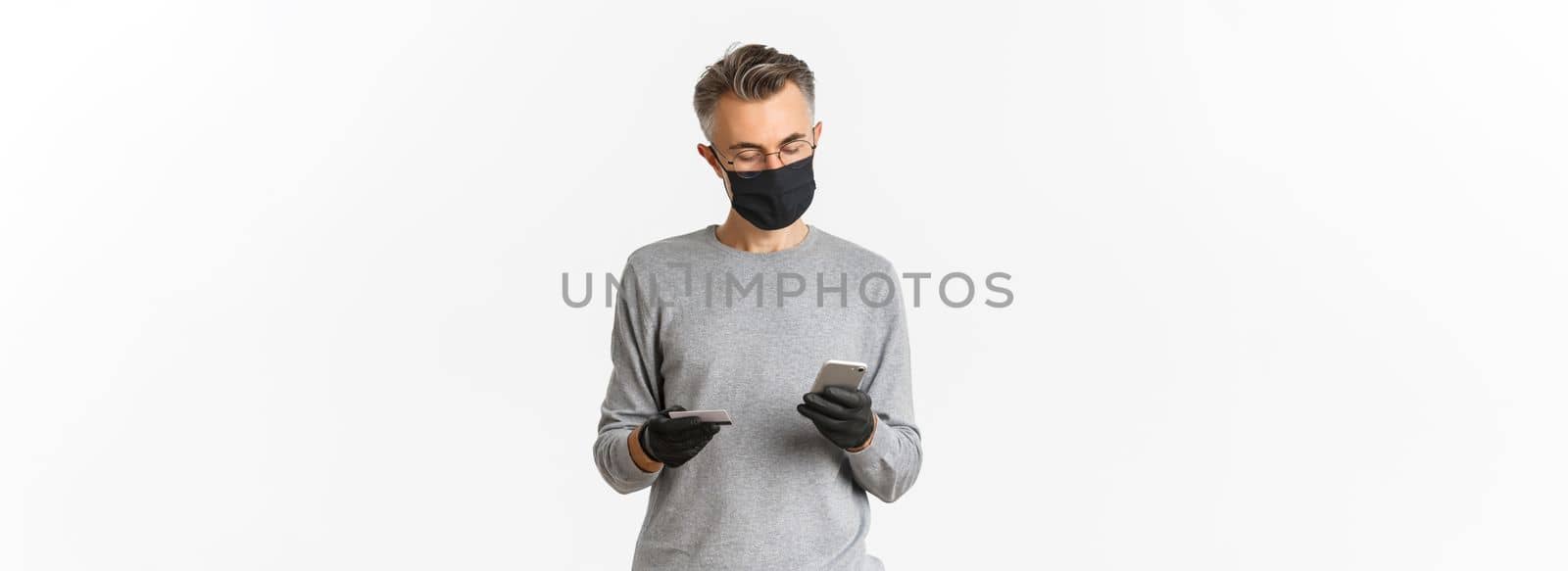 Concept of covid-19, social distancing and lifestyle. Image of handsome middle-aged man in medical mask, gloves and glasses, making online order using credit card and mobile phone.