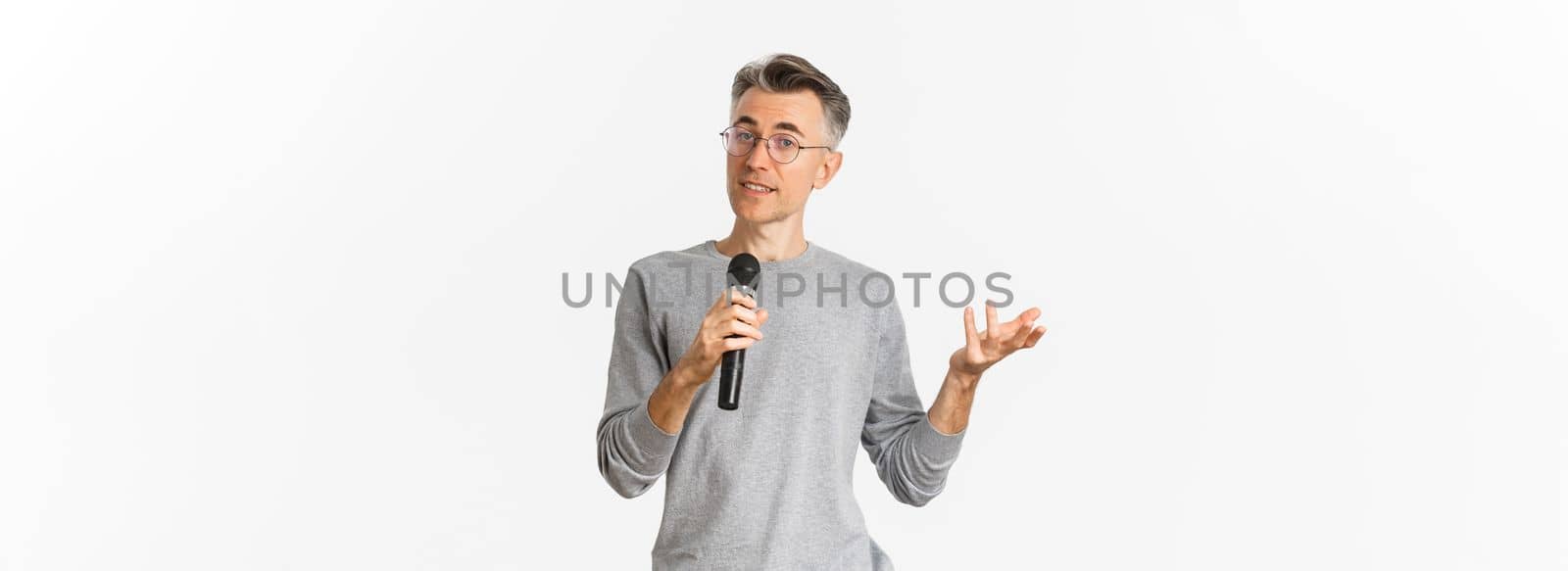 Portrait of handsome middle-aged man in glasses and gray sweater, making speech, talking with microphone and explaining something, standing over white background.