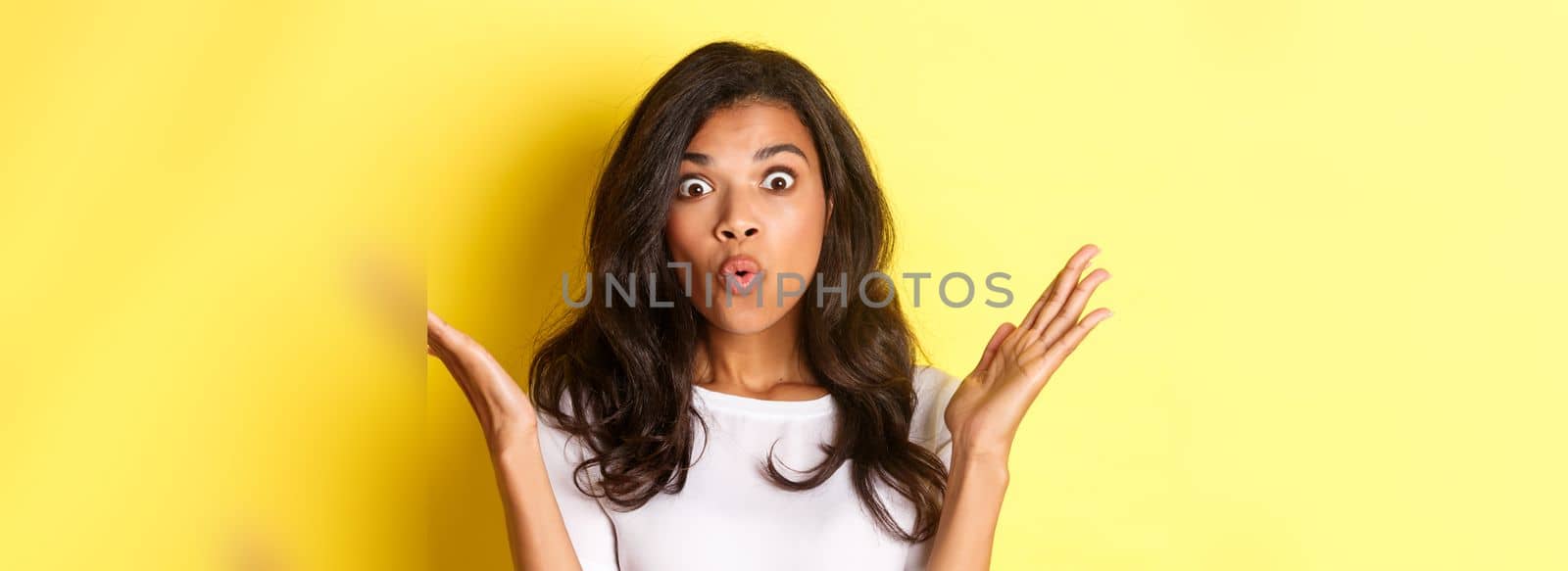 Close-up of surprised african-american girl, raising hands up and saying wow, hear amazing news, standing over yellow background.