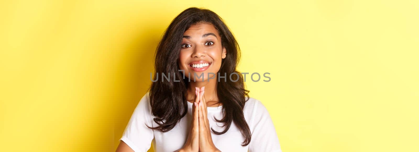 Close-up of cheerful african-american girl, smiling and saying thank you, press hands over chest in pray gesture, standing over yellow background.