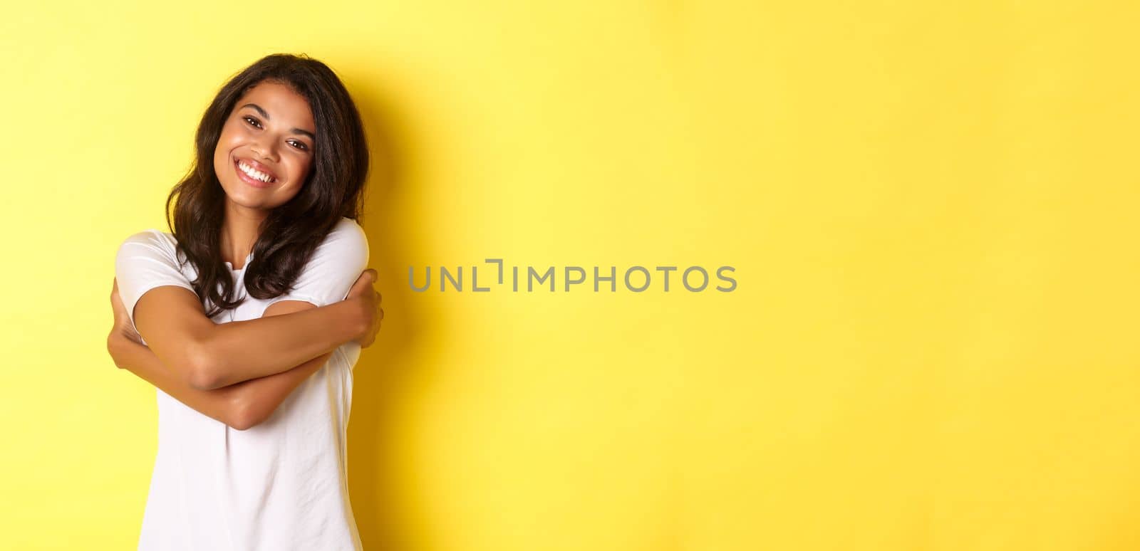 Portrait of cheerful african american woman, hugging herself and smiling pleased, standing over yellow background.