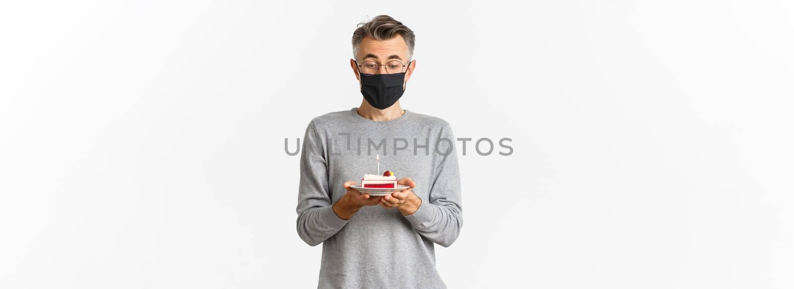 Concept of covid-19, quarantine and holidays. Image of dreamy middle-aged man in medical mask and glasses, looking amazed at birthday cake, making a wish, standing over white background by Benzoix