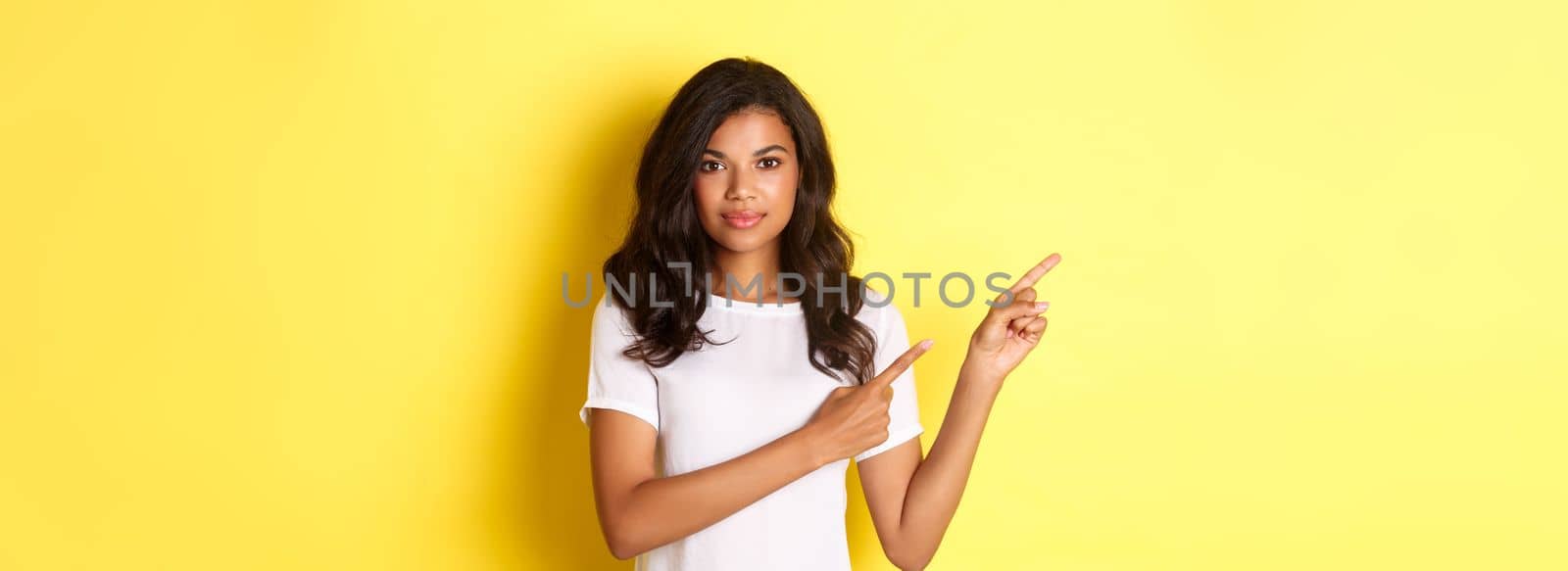 Portrait of beautiful and confident african-american woman, pointing fingers left at copy space, showing logo banner and smiling, standing over yellow background.