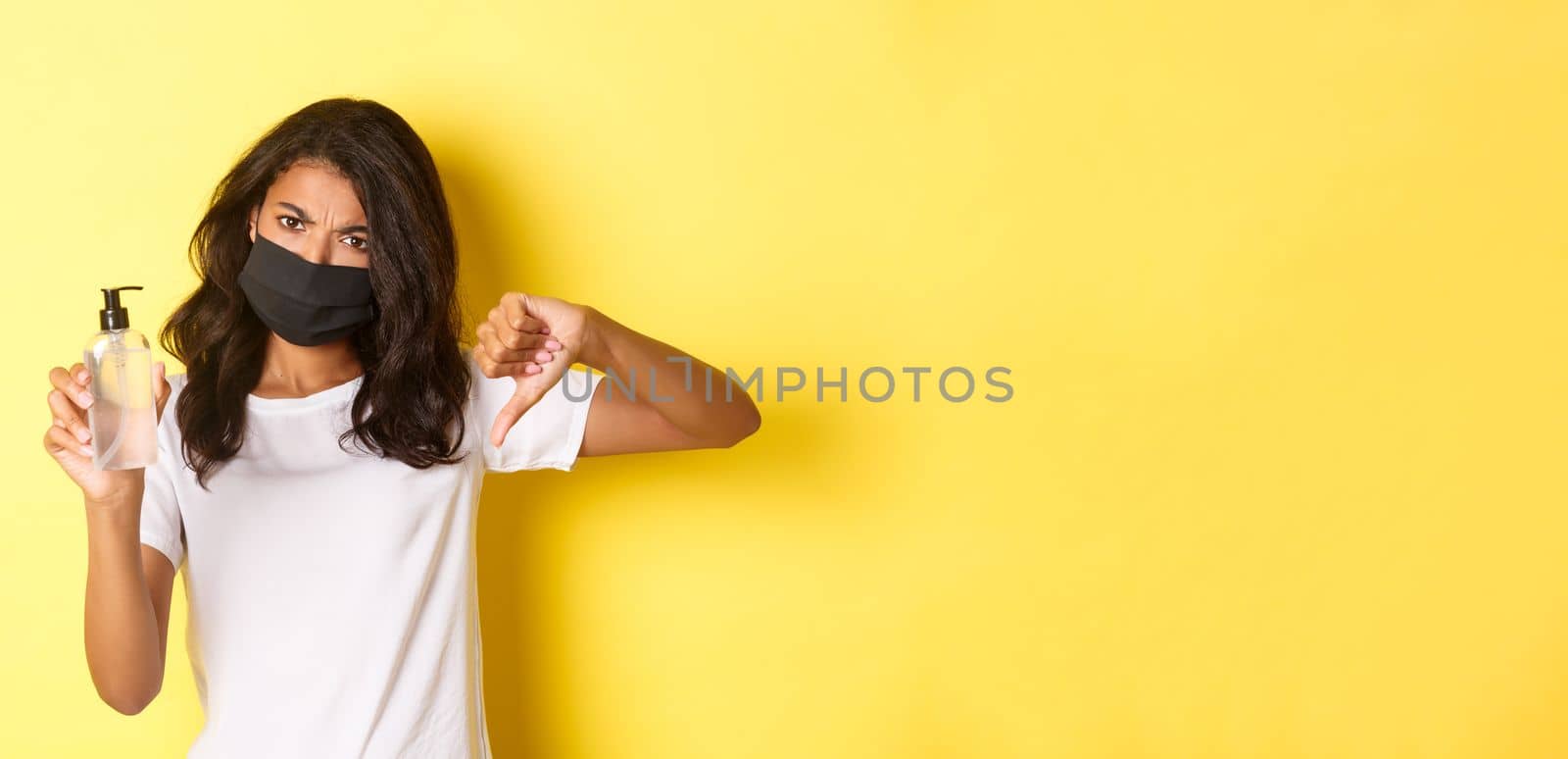 Concept of covid-19, social distancing and lifestyle. Image of disappointed african-american woman in face mask, showing thumbs-down, do not recommend bad hand sanitizer, yellow background by Benzoix