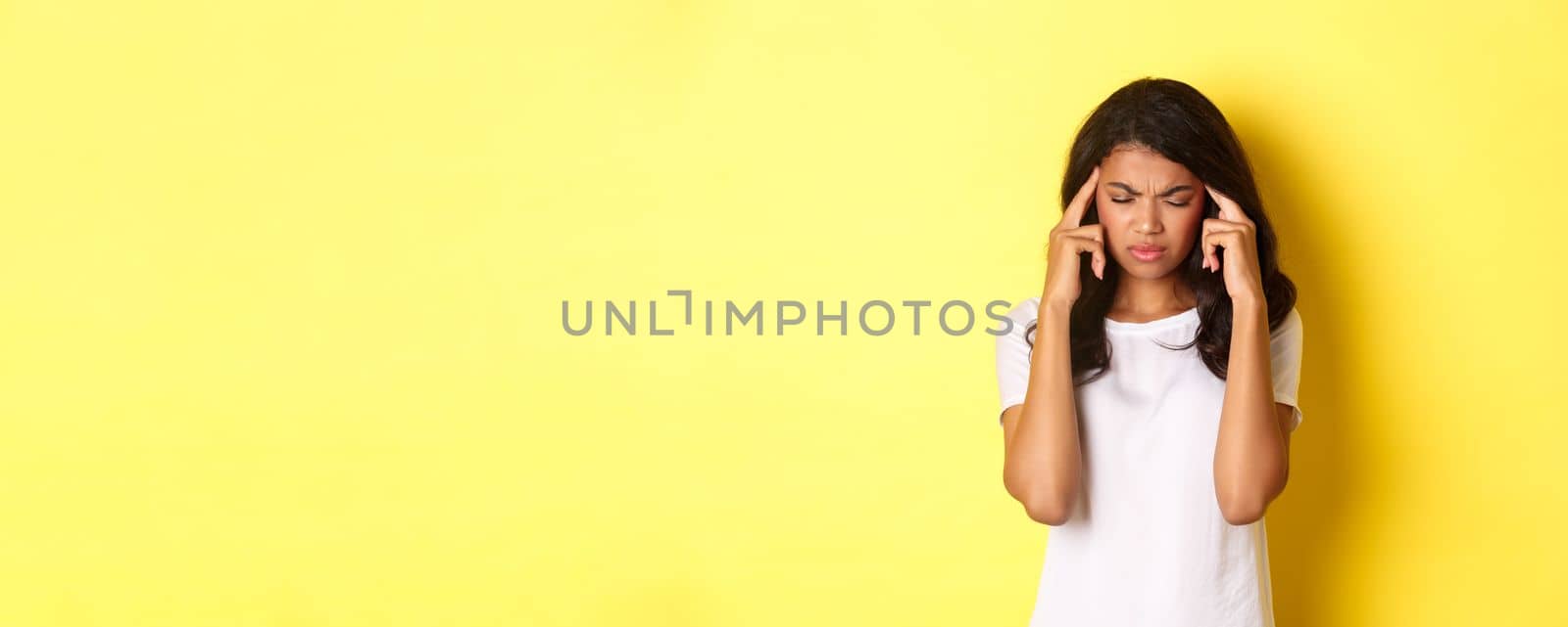 Portrait of young african-american female model feeling sick, frowning and touching head, complaining on headache, standing over yellow background.