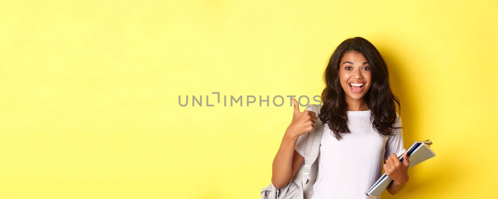 Image of cheerful african-american girl student, holding notebooks for classes and backpack, showing thumbs-up, like studying, standing over yellow background.