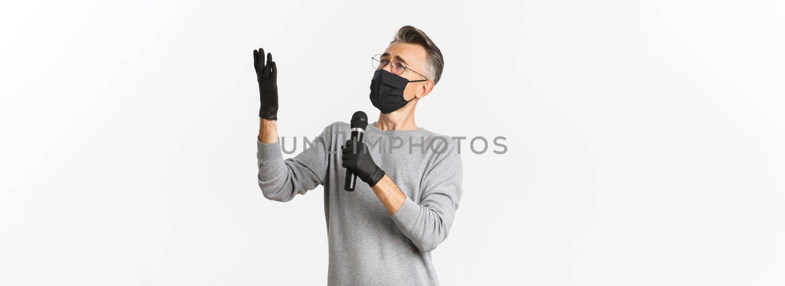 Concept of coronavirus, lifestyle and quarantine. Portrait of charismatic middle-aged man in medical mask and gloves, singing serenade in microphone, standing over white background.