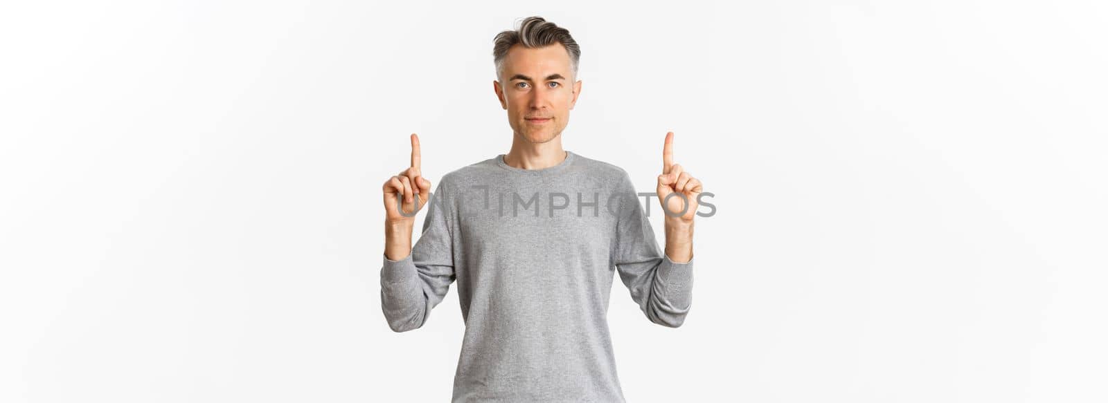 Portrait of handsome middle-aged man in grey sweater, showing promo, pointing fingers up and looking serious, standing over white background.