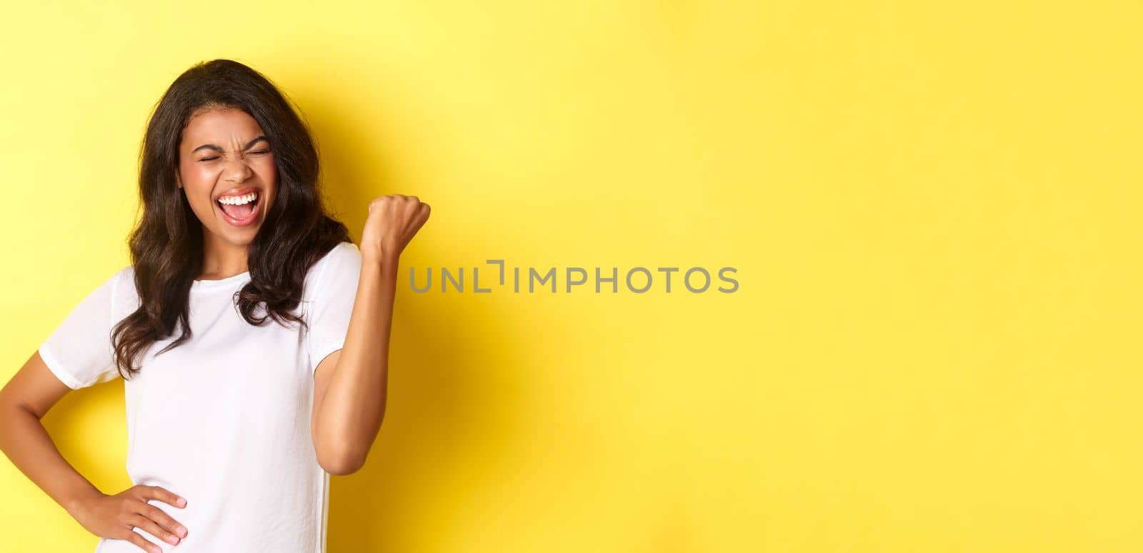 Portrait of happy african-american woman, triumphing and winning, making fist pump and shouting for joy, standing pleased against yellow background by Benzoix