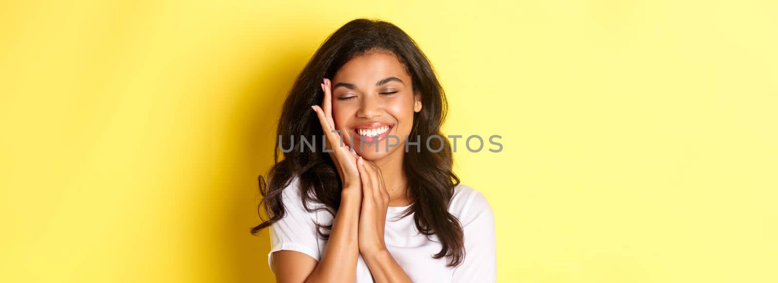 Close-up of dreamy, beautiful african-american woman, gently touching face and smiling pleased, standing against yellow background.