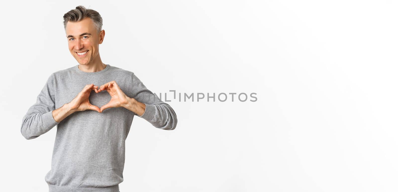 Portrait of cheeky middle-aged man in grey sweater, smiling and showing heart sign, love someone, standing over white background.
