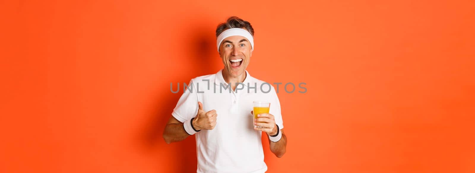 Concept of workout, gym and lifestyle. Image of active and healthy middle-aged sport guy, showing thumbs-up and drinking juice, smiling happy, standing over orange background.