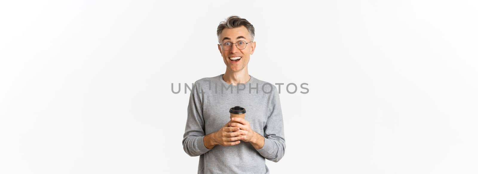 Portrait of surprised and happy middle-aged man in glasses and grey sweater, drinking coffee from takeaway cup and looking amazed, standing over white background.