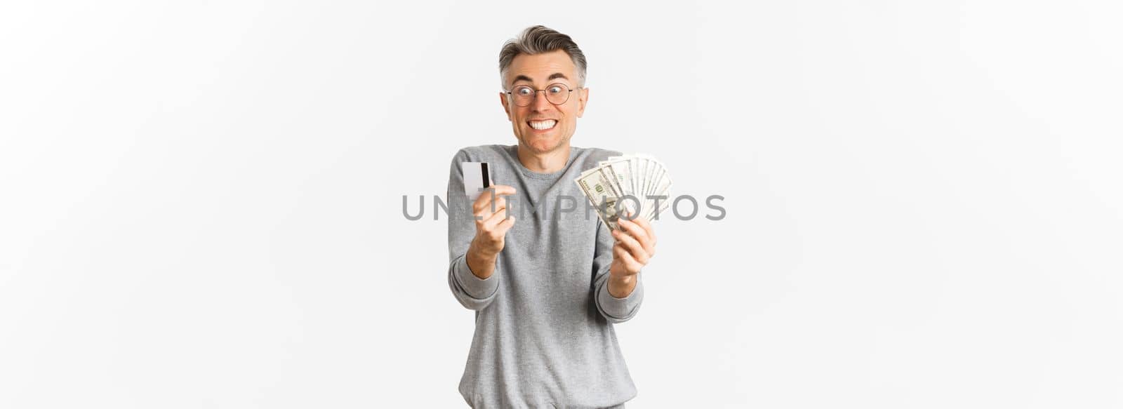 Portrait of excited and happy middle-aged man, looking amazed at credit card and money, standing over white background.