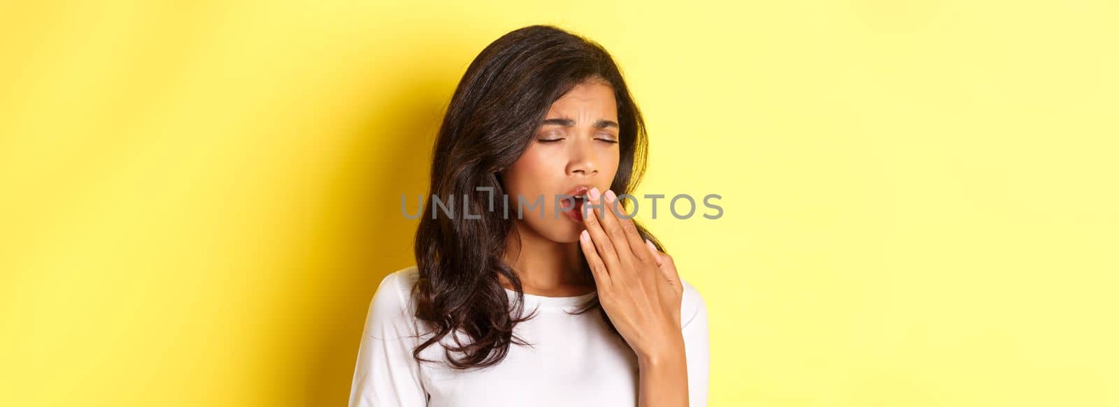 Close-up of tired and sleepy african-american girl, yawning and looking exhausted, standing in white t-shirt over yellow background.