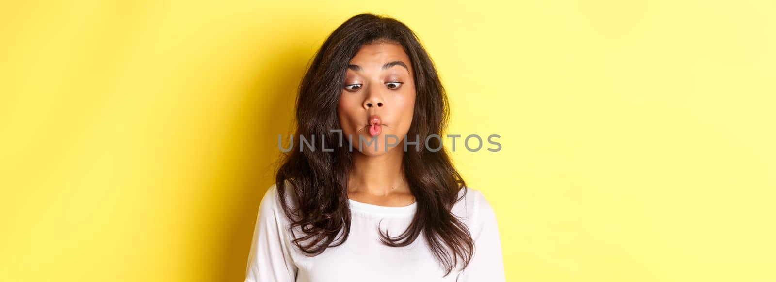 Close-up of funny african-american woman, making faces, squinting and pouting, fooling around over yellow background.