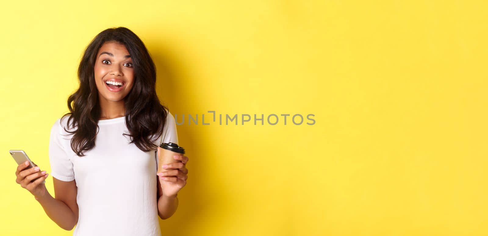 Portrait of attractive african-american girl smiling, holding coffee cup and smartphone, standing over yellow background by Benzoix