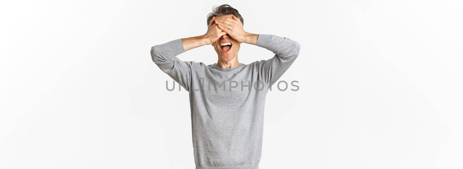 Portrait of happy middle-aged man waiting for surprise, shut eyes and smiling, playing hide n seek, standing over white background.
