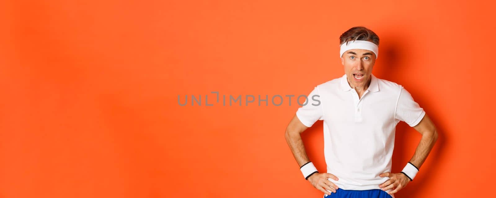 Concept of workout, sports and lifestyle. Portrait of startled middle-aged fitness guy in activewear, drop jaw and looking at camera amazed, standing over orange background.