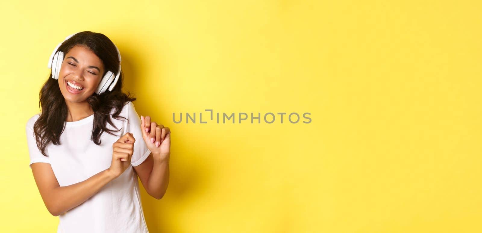 Cheerful teenage african-american girl listening to music in headphones, dancing upbeat and smiling, standing over yellow background.
