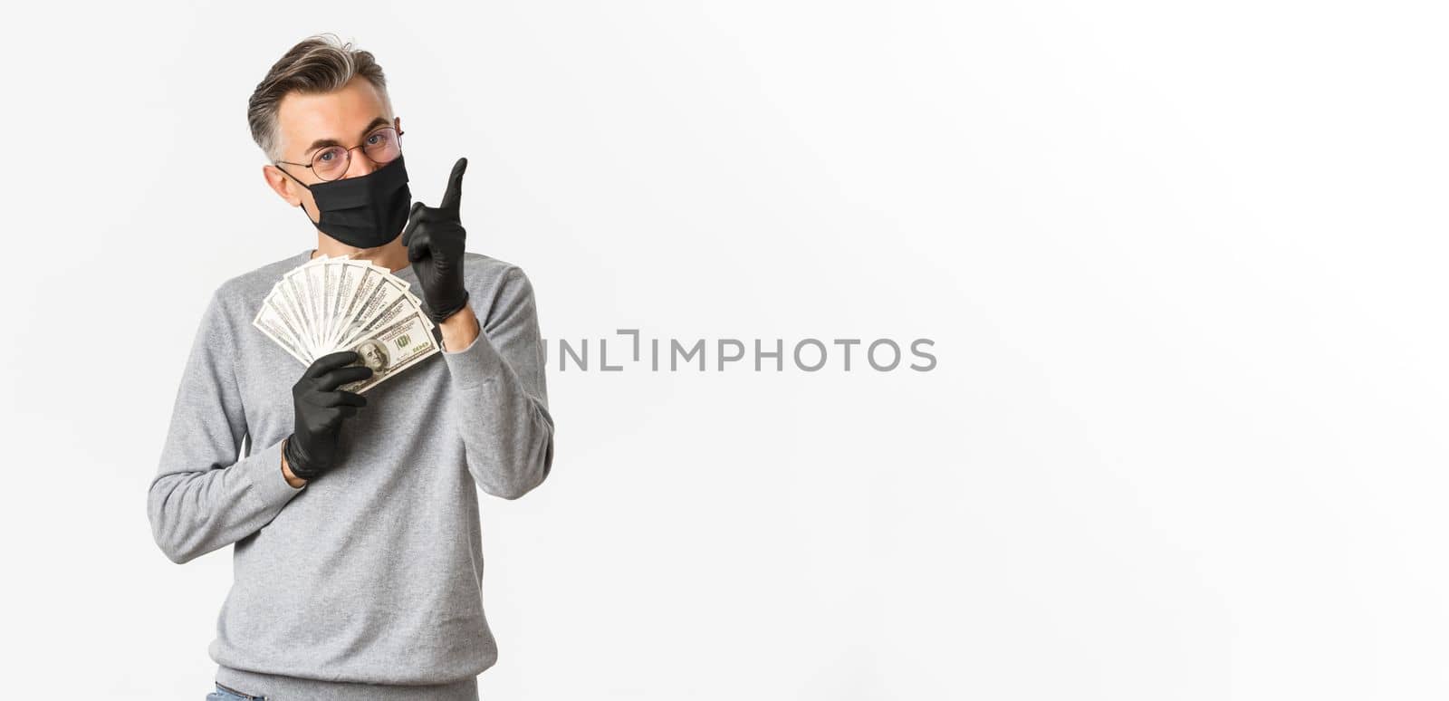 Concept of covid-19, social distancing and lifestyle. Portrait of lucky middle-aged man in medical mask, gloves and glasses, showing money and pointing finger up, smiling pleased by Benzoix