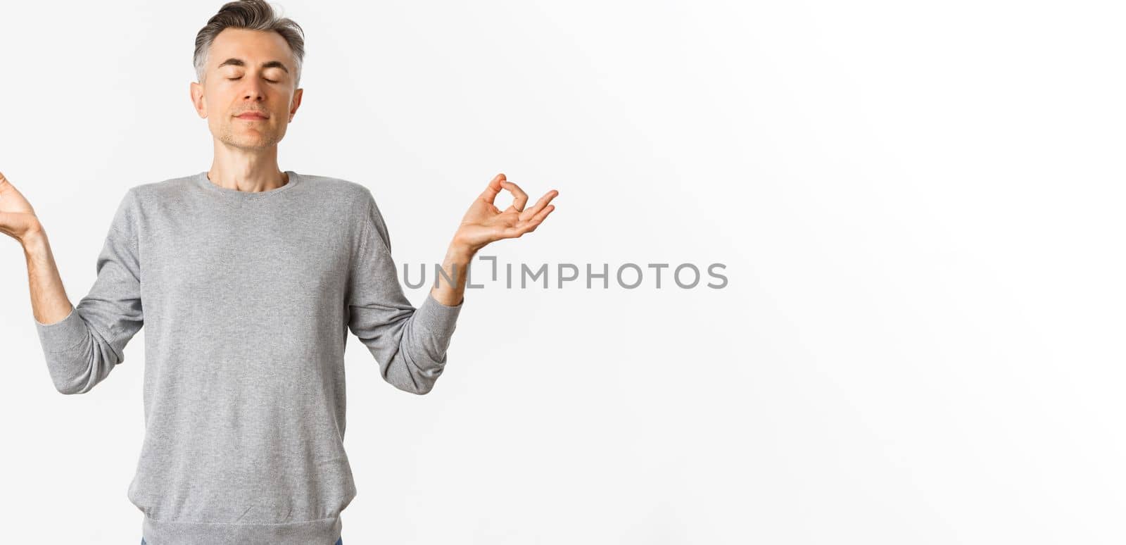 Portrait of calm and relaxed middle-aged man, breathing air freely, meditating with eyes closed and hands spread sideways, practice yoga over white background.