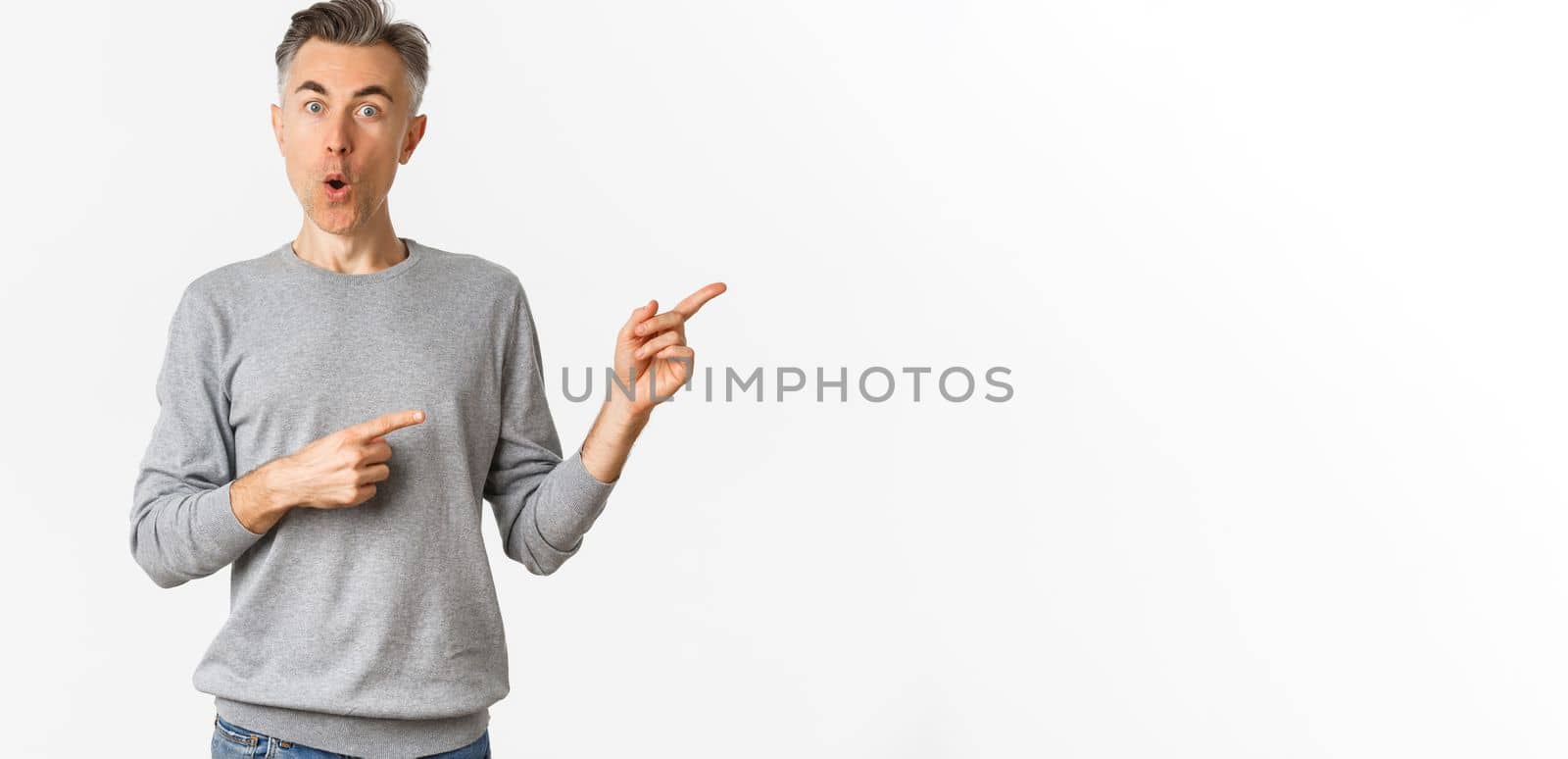 Amazed handsome guy in grey sweater, pointing fingers right and looking curious, askign question about product, standing over white background.