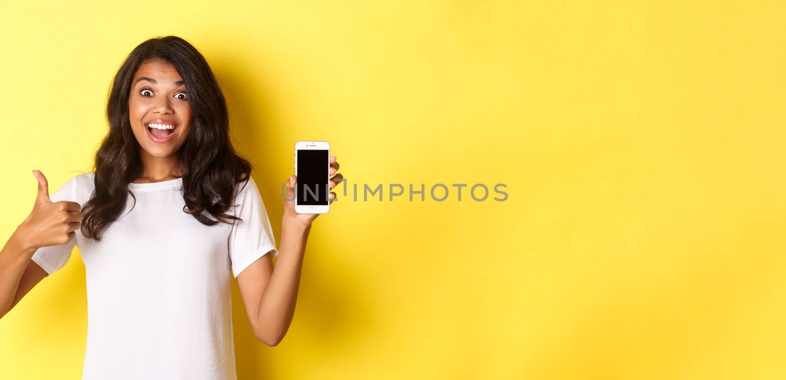 Portrait of excited african-american girl, showing smartphone screen and making thumbs-up, recommending something cool, standing over yellow background by Benzoix