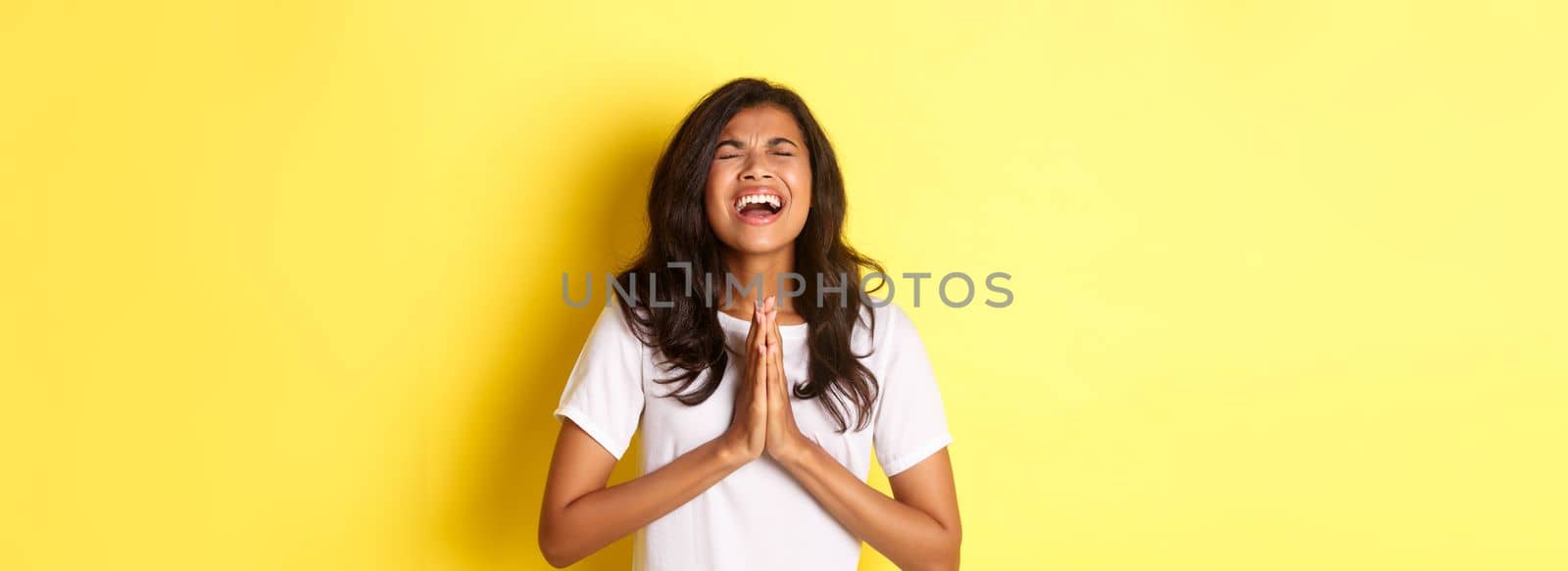 Portrait of young african-american girl pleading, begging for help with hands pressed together on chest, standing in white t-shirt over yellow background by Benzoix