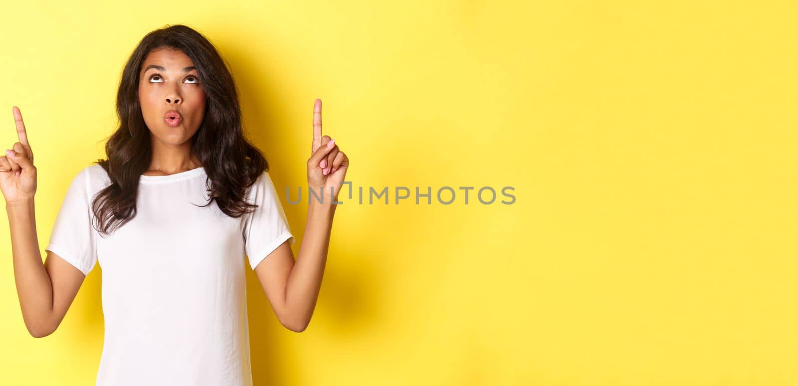 Portrait of amazed and surprised african-american girl, saying wow, pointing and looking up at cool offer banner, standing over yellow background.