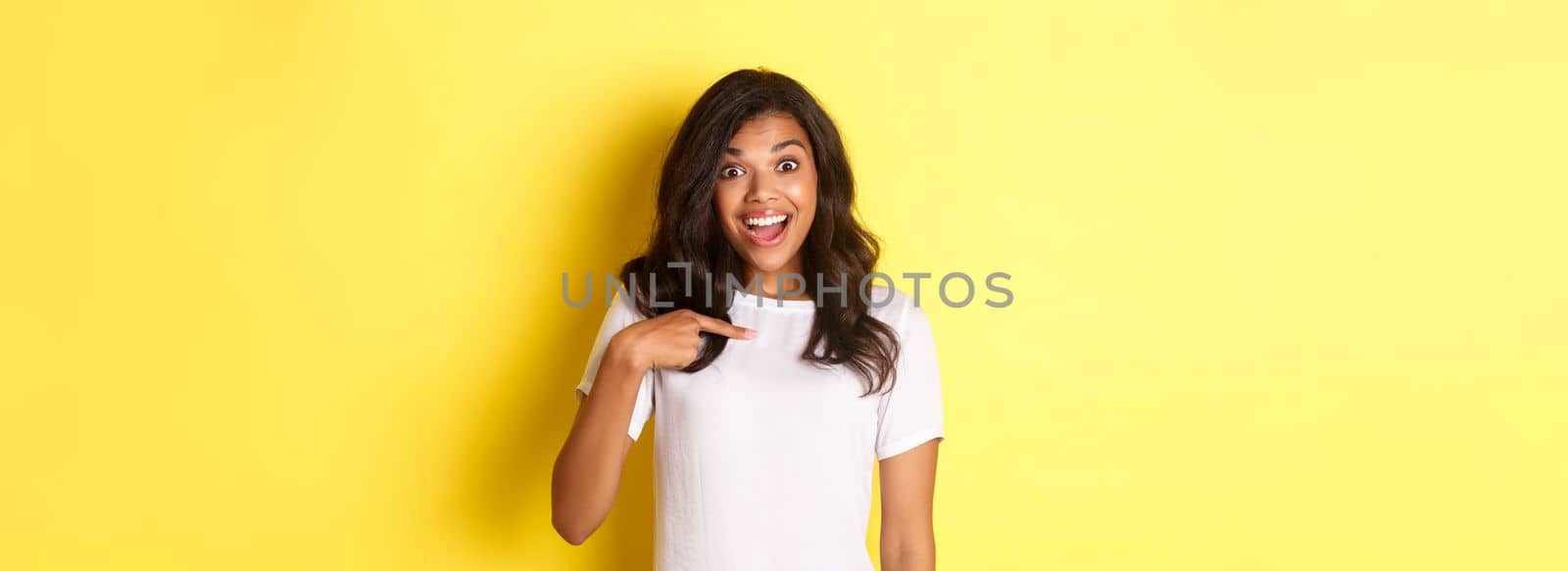 Image of attractive african-american girl looking amazed and happy, smiling while pointing at herself, being chosen, standing over yellow background.