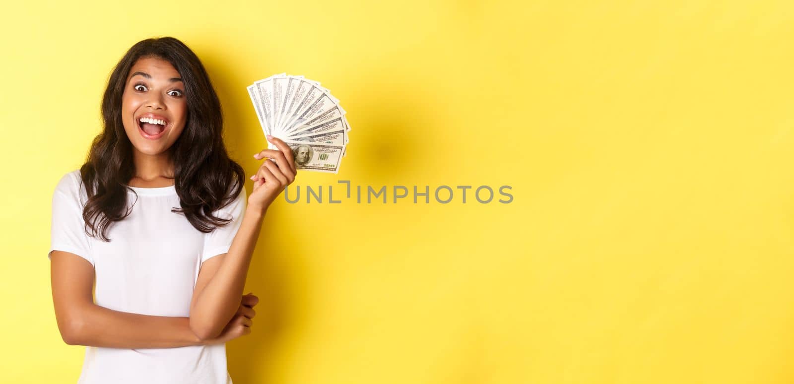 Portrait of beautiful african-american girl, smiling happy and showing money, going shopping, standing over yellow background.