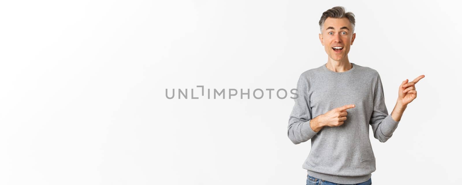 Image of surprised handsome middle-aged man, smiling and pointing fingers right at logo or promo banner, standing amazed over white background.