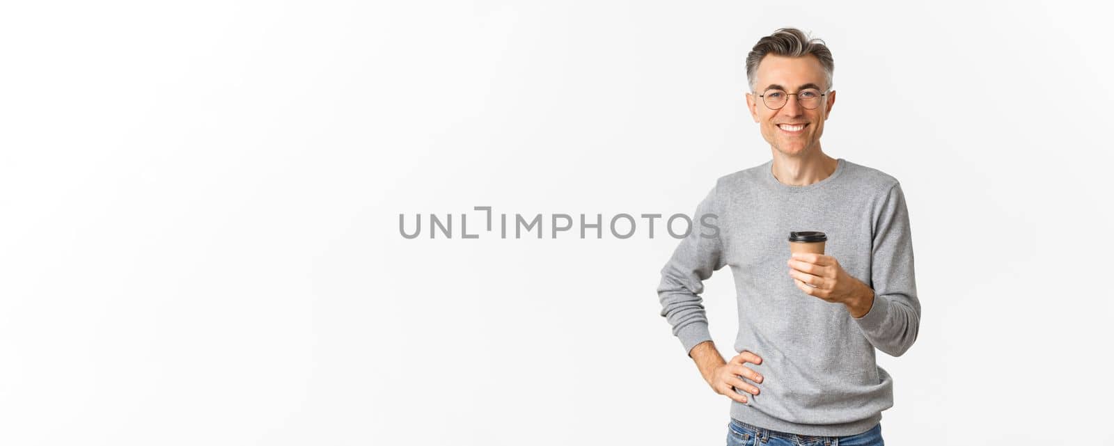 Image of confident and handsome middle aged man, wearing glasses and grey sweater, smiling happy and drinking coffee from takeaway cup, standing satisfied over white background.
