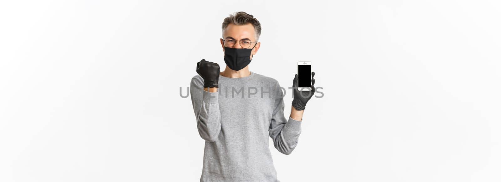 Concept of covid-19, social distancing and lifestyle. Happy middle-aged man in medical mask, gloves and glasses rejoicing, showing achievement on smartphone screen.