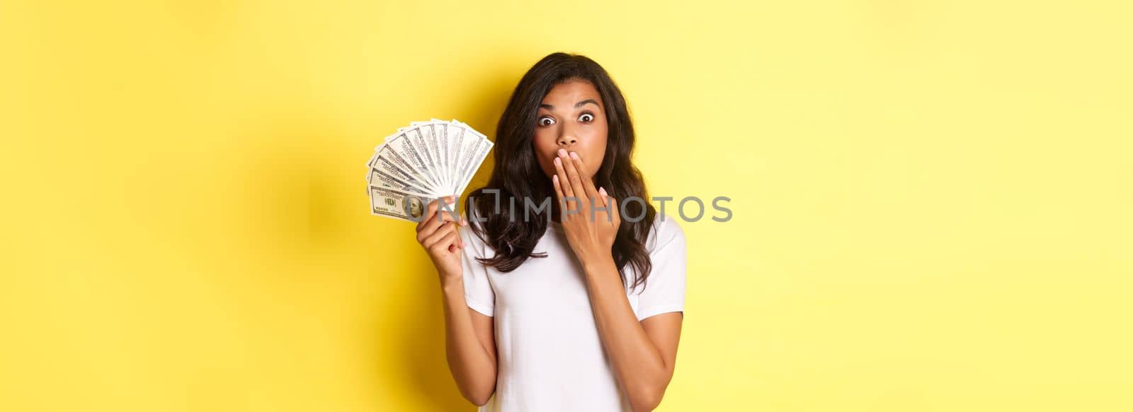 Image of surprised african-american girl winning money, gasping amazed, standing over yellow background by Benzoix